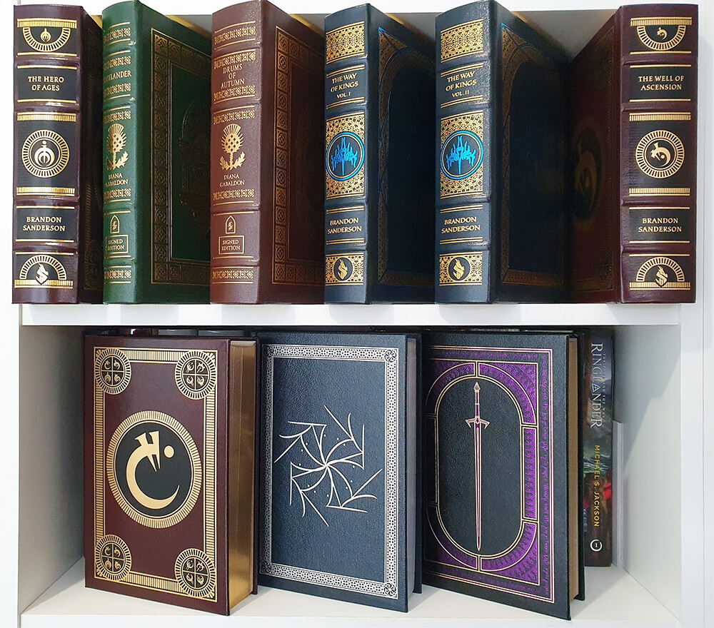 Nine leather-bound books on bookshelves. The books are in different colour leathers -- some brown, some green, some dark blue. They all have gilding of some kind and designs of varying intricacy. Some have the titles visible: there's the Mistborn trilogy, The Way of Kings by Brandon Sanderson, Outlander, and Drums of Autumn.