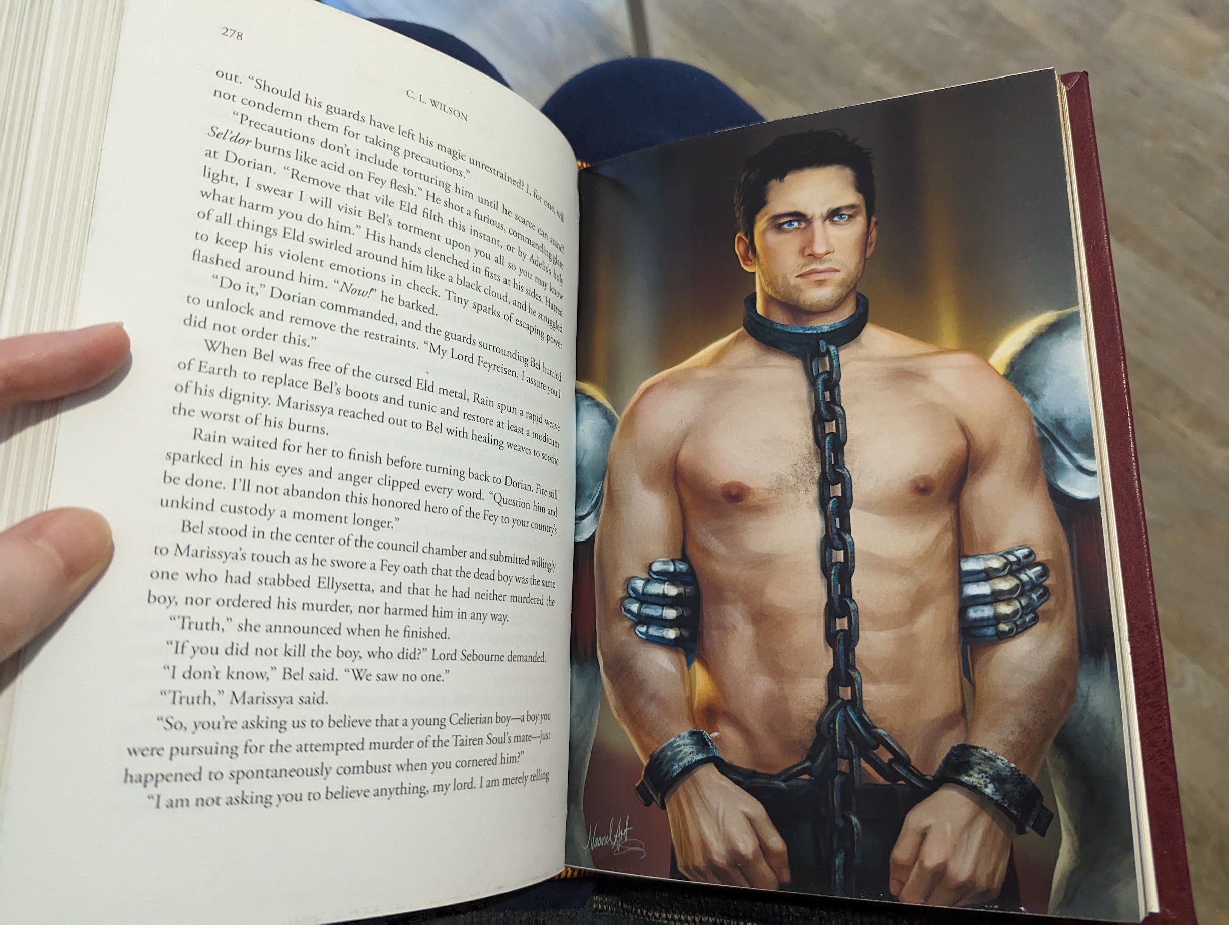 Photo of a full colour illustration inside a book. It features a muscular, shirtless man with a chain around his neck and shackles around his wrists.