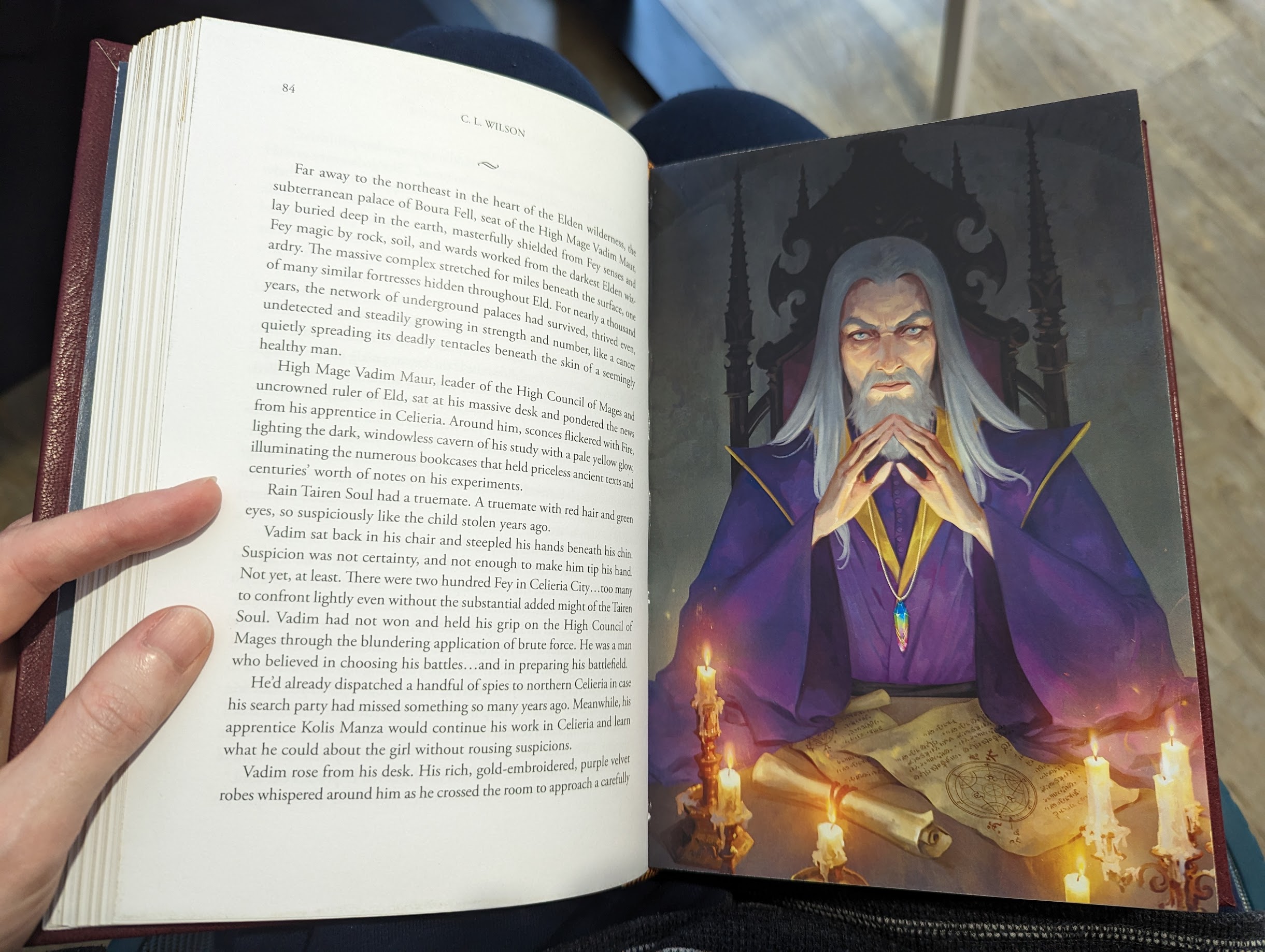 Photo of a full colour illustration inside a book. It depicts High Mage Vadim Maur from Tairen Soul. An evil looking mage with grey/white hair and purple robes. He's sitting at a desk containing various scrolls and candles.