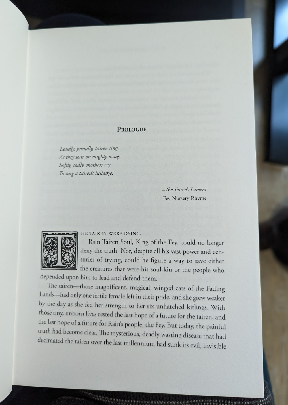 Photo of the page of a book. At the top it says "Prologue" in large heading text, with a short poem underneath it. The first paragraph has a drop cap (letter "T" with intricate flower designs around it).
